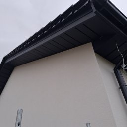 Piotr Mroczek REM_BLACH - Czarny dach z rynnami i podbitką w Strumieniu, widziany z dołu. Fragment rusztowania w kadrze. Elewacja budynku w jasnym kolorze. Pochmurne niebo w tle.