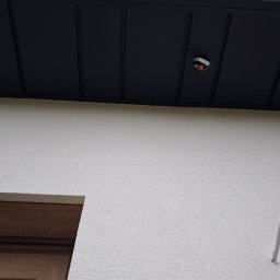 Piotr Mroczek REM_BLACH - Czarny podbitka dachowa z widocznym punktem oświetleniowym na tle białej, teksturowanej ściany budynku i fragmentu okna z drewnianą ramą. Ujęcie z dołu.