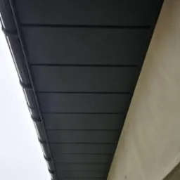 Nowe, ciemnoszare pokrycie dachowe z zamontowanym systemem rynnowym, widoczne z dołu pod kątem, z fragmentem ściany budynku w tle.