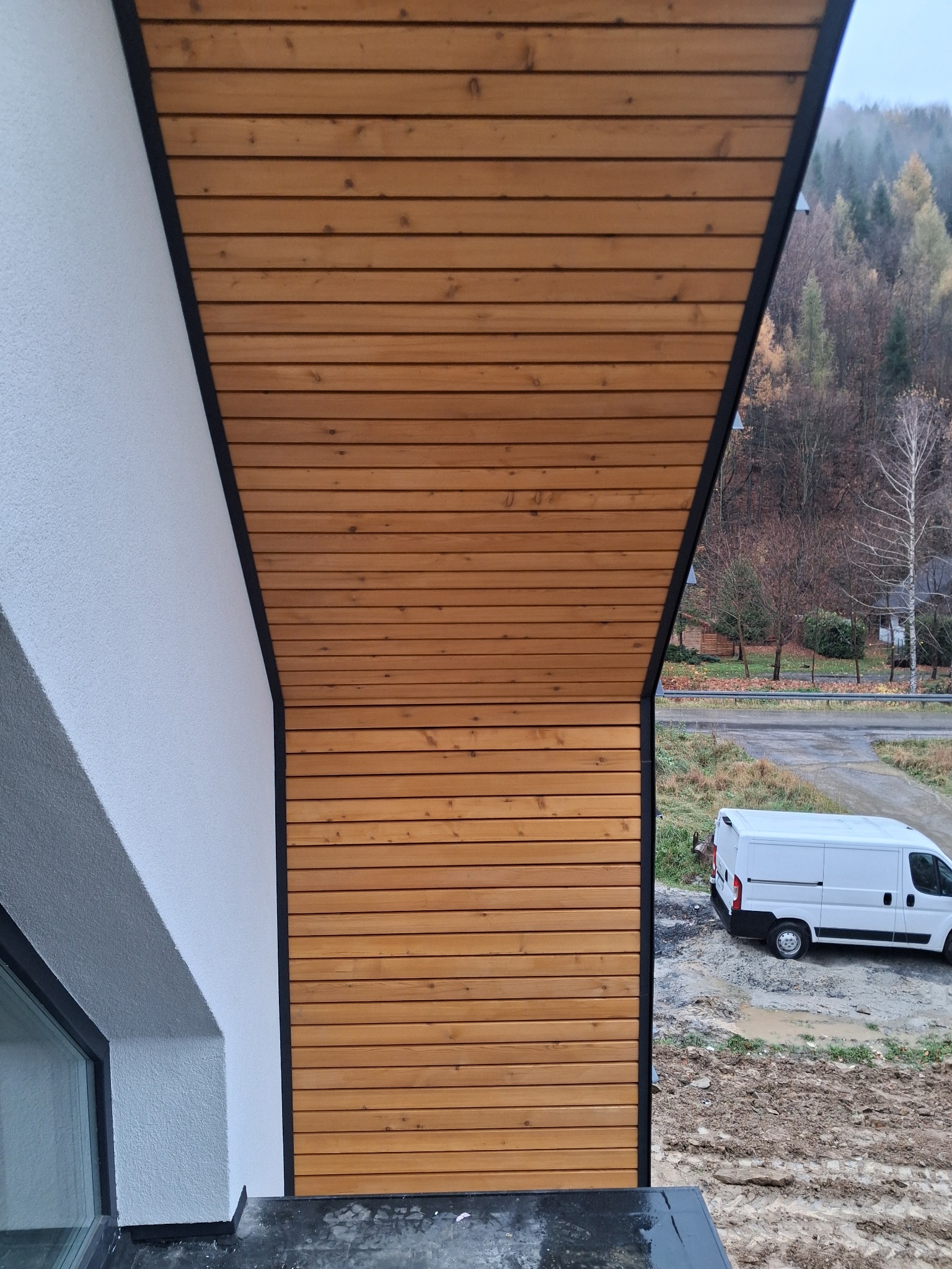 Drewniana elewacja budynku z czarnymi obramowaniami, widok z góry, w tle zalesione wzgórza i biały samochód dostawczy. Detal wykończenia zewnętrznego.