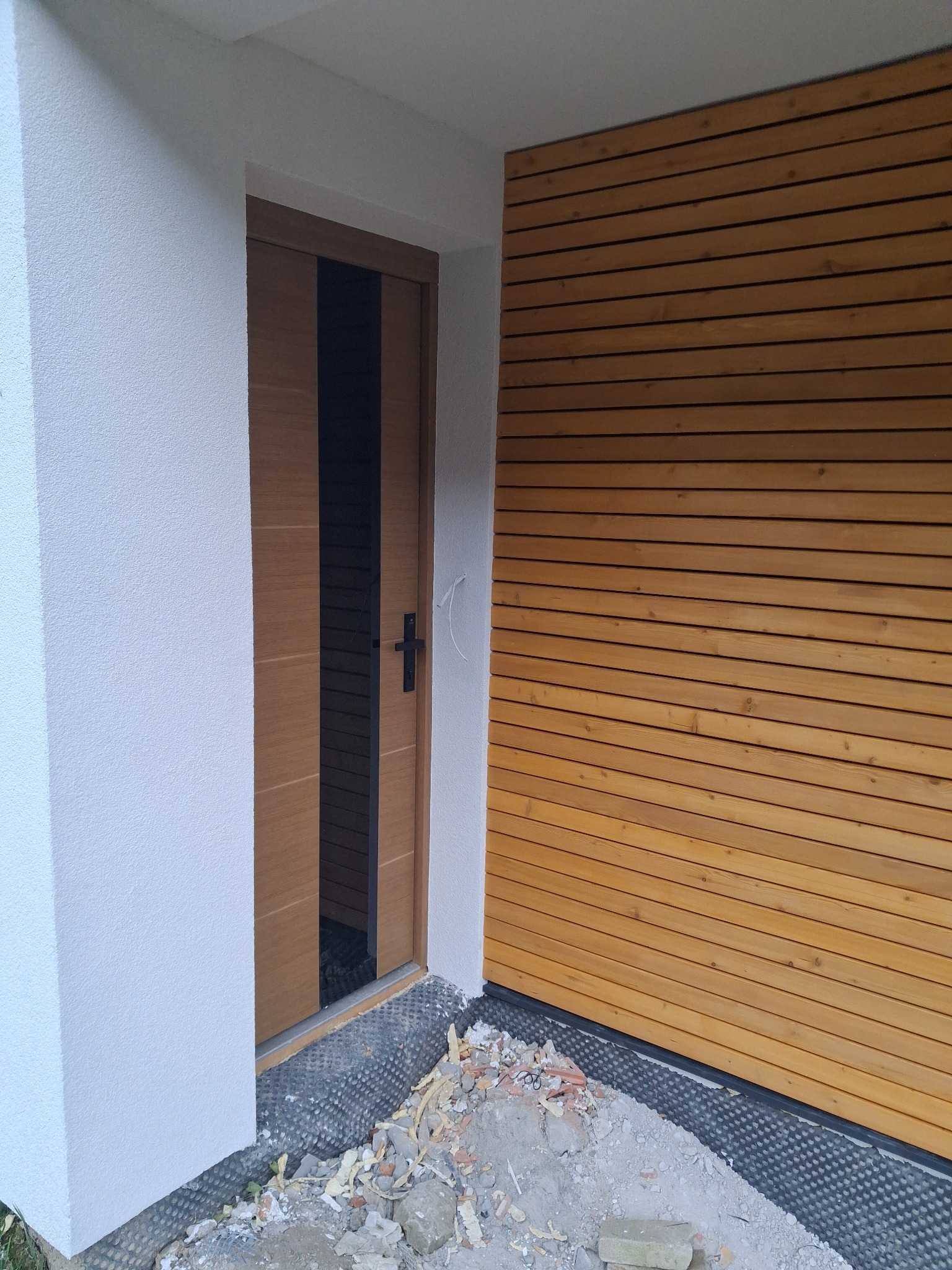 Nowoczesne wykończenie elewacji z drewnianą okładziną i otwartymi drzwiami. Widoczne detale montażu i pozostałości po pracach budowlanych.