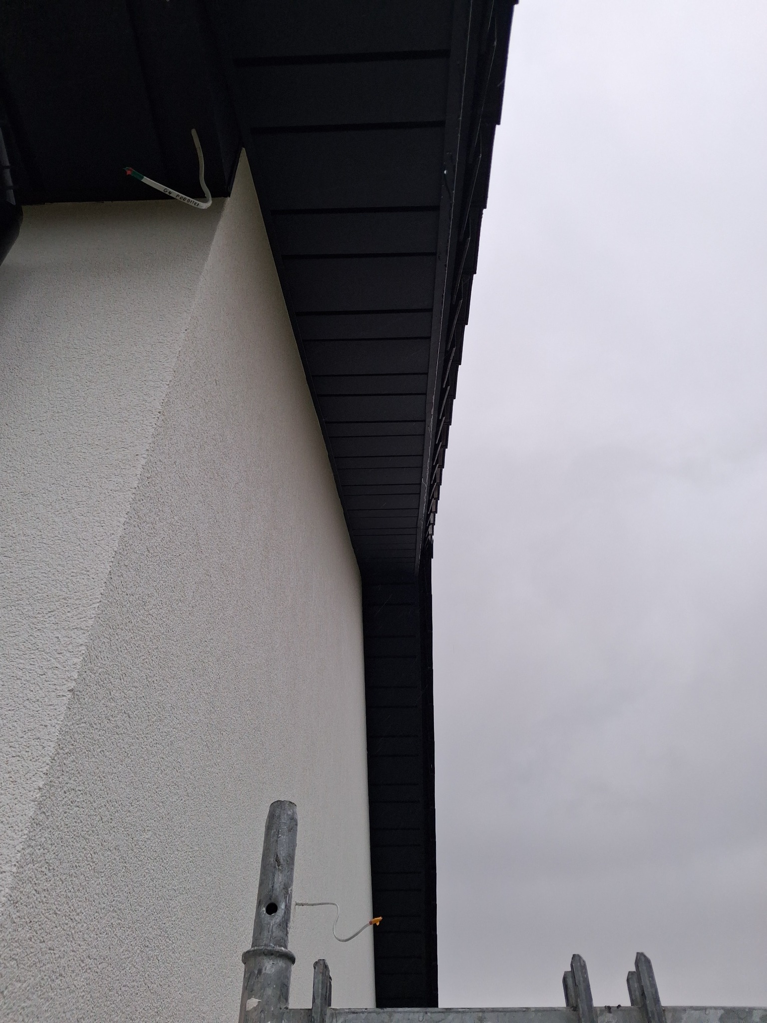 Fragment elewacji budynku z widoczną podbitką dachową i fragmentem rusztowania. Ściana o jasnej, ziarnistej fakturze, podbitka w kolorze ciemnego brązu. Pochmurne niebo w tle.