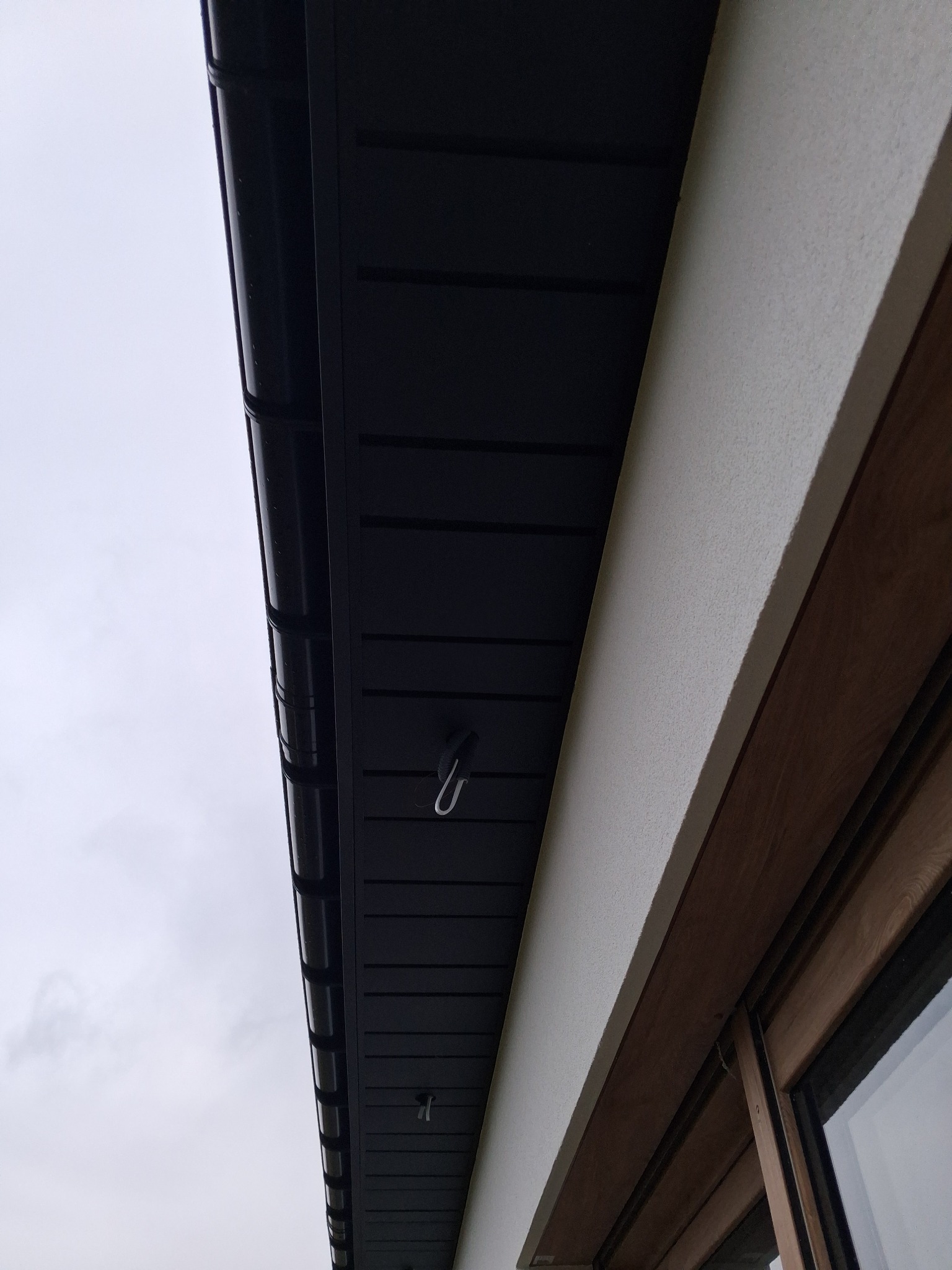 Czarne podbitki dachowe z widocznymi hakami, zamontowane przy białej ścianie budynku z oknem. Ujęcie z dołu, pochmurne niebo w tle.