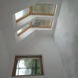 Poddasze z dwoma oknami dachowymi i jednym pionowym, ściany w trakcie przygotowania do malowania, widoczne ślady szpachlowania i gładzi.