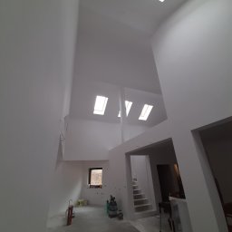 B&F Kompleksowe Wykończenia Wnętrz - Wnętrze dwupoziomowego pomieszczenia z wysokim sufitem i oknami dachowymi po świeżym malowaniu na biało, widoczne ślady po pracach wykończeniowych, narzędzia i farby w tle.