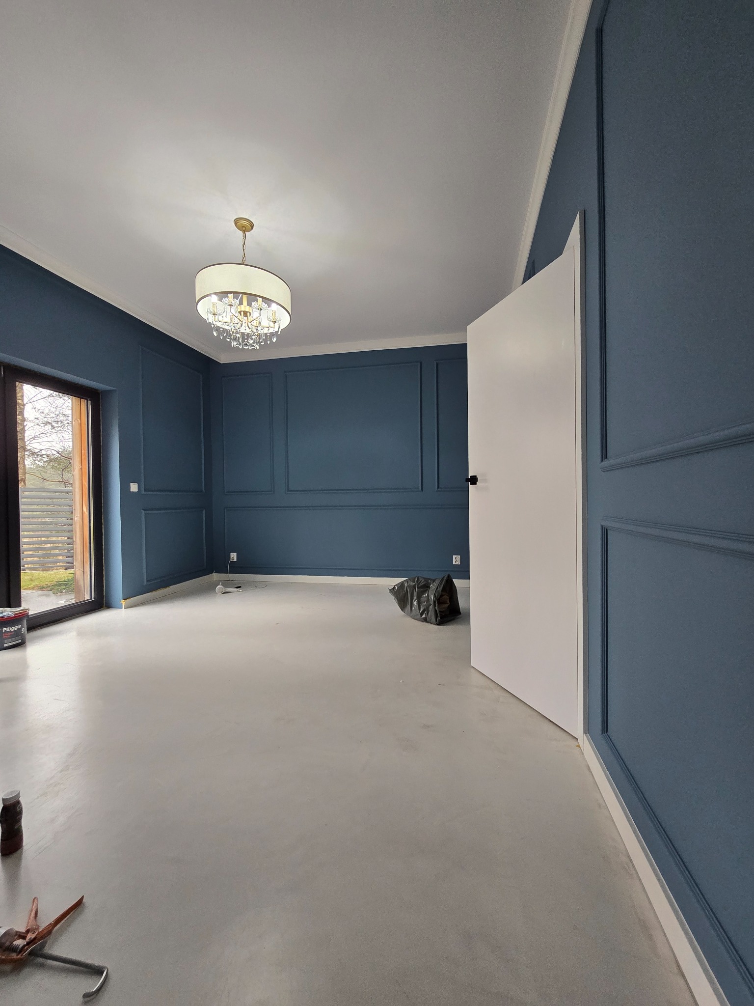 Pokój po remoncie: granatowe ściany z dekoracyjnymi listwami, biały sufit z żyrandolem i jasna podłoga. Widoczne narzędzia i otwarte drzwi balkonowe.