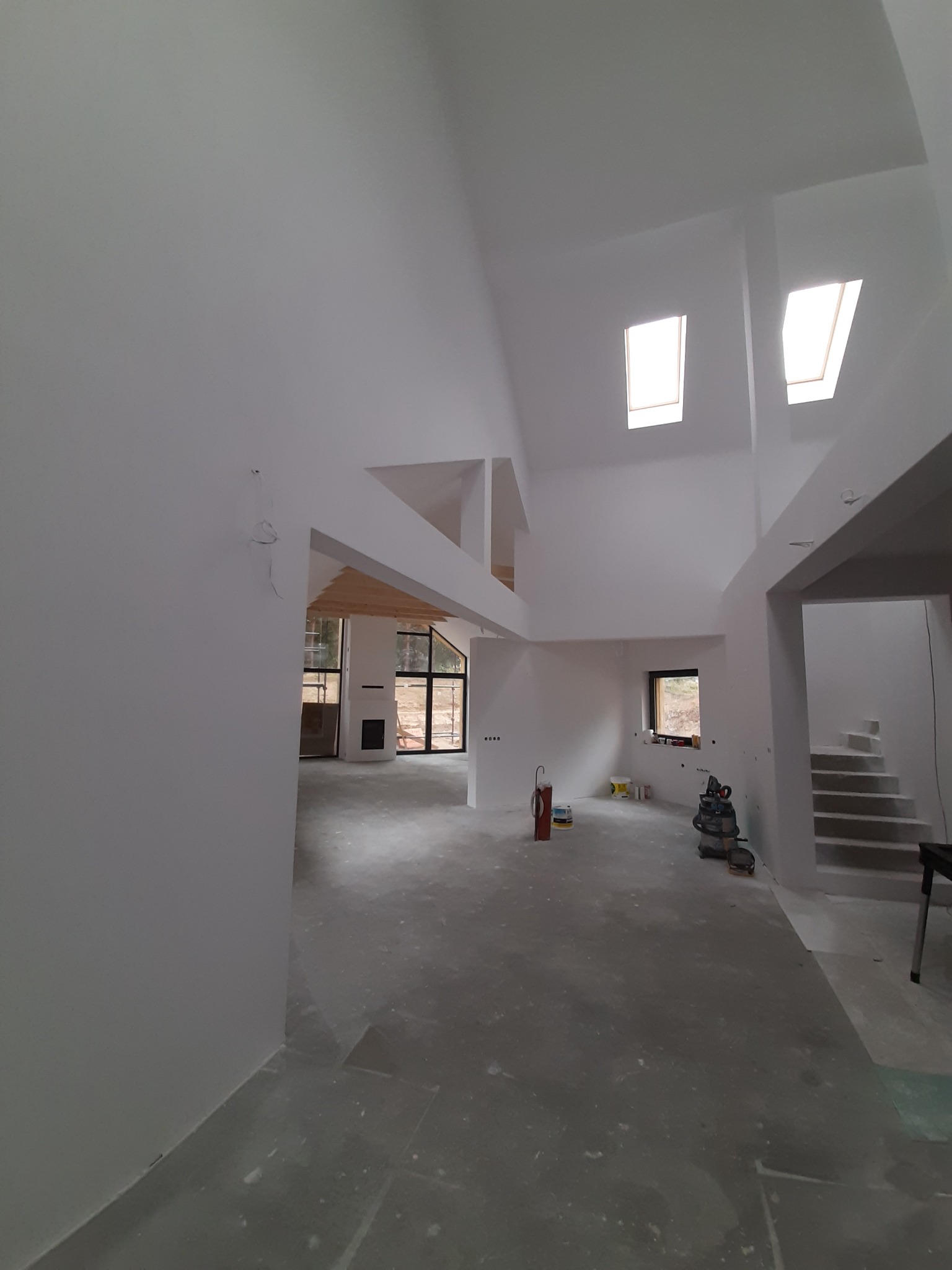 Wnętrze domu w trakcie prac wykończeniowych, ściany pomalowane na biało, surowa podłoga, widoczne okna dachowe i sprzęt budowlany.