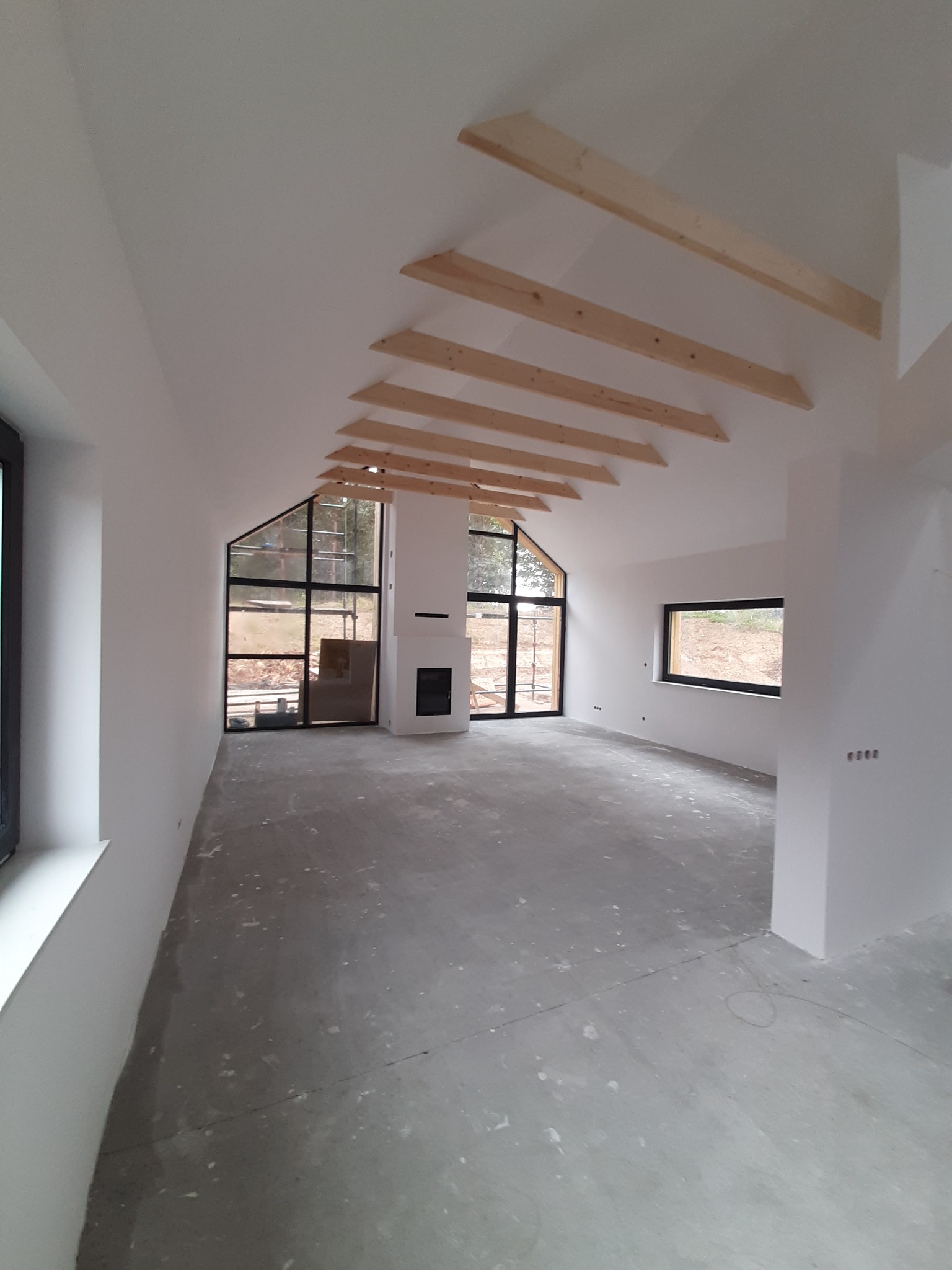 Surowe wnętrze w trakcie wykończenia: białe ściany, surowy beton na podłodze, drewniane belki stropowe, duże okna z czarnymi ramami, widok na zewnątrz na plac budowy.