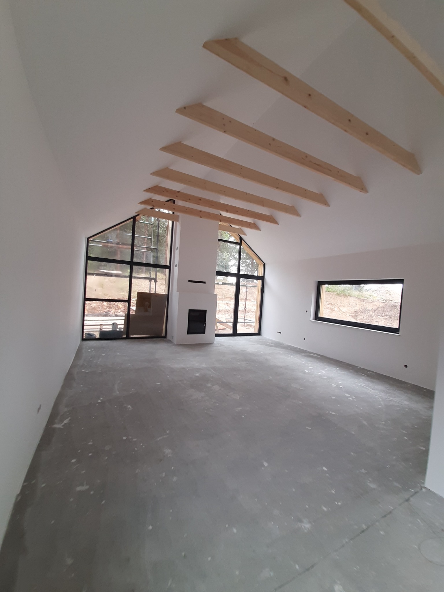 Wnętrze poddasza z surowymi belkami stropowymi, dużymi oknami w czarnych ramach i kominkiem, ściany pomalowane na biało, surowa wylewka na podłodze, w trakcie prac wykończeniowych.