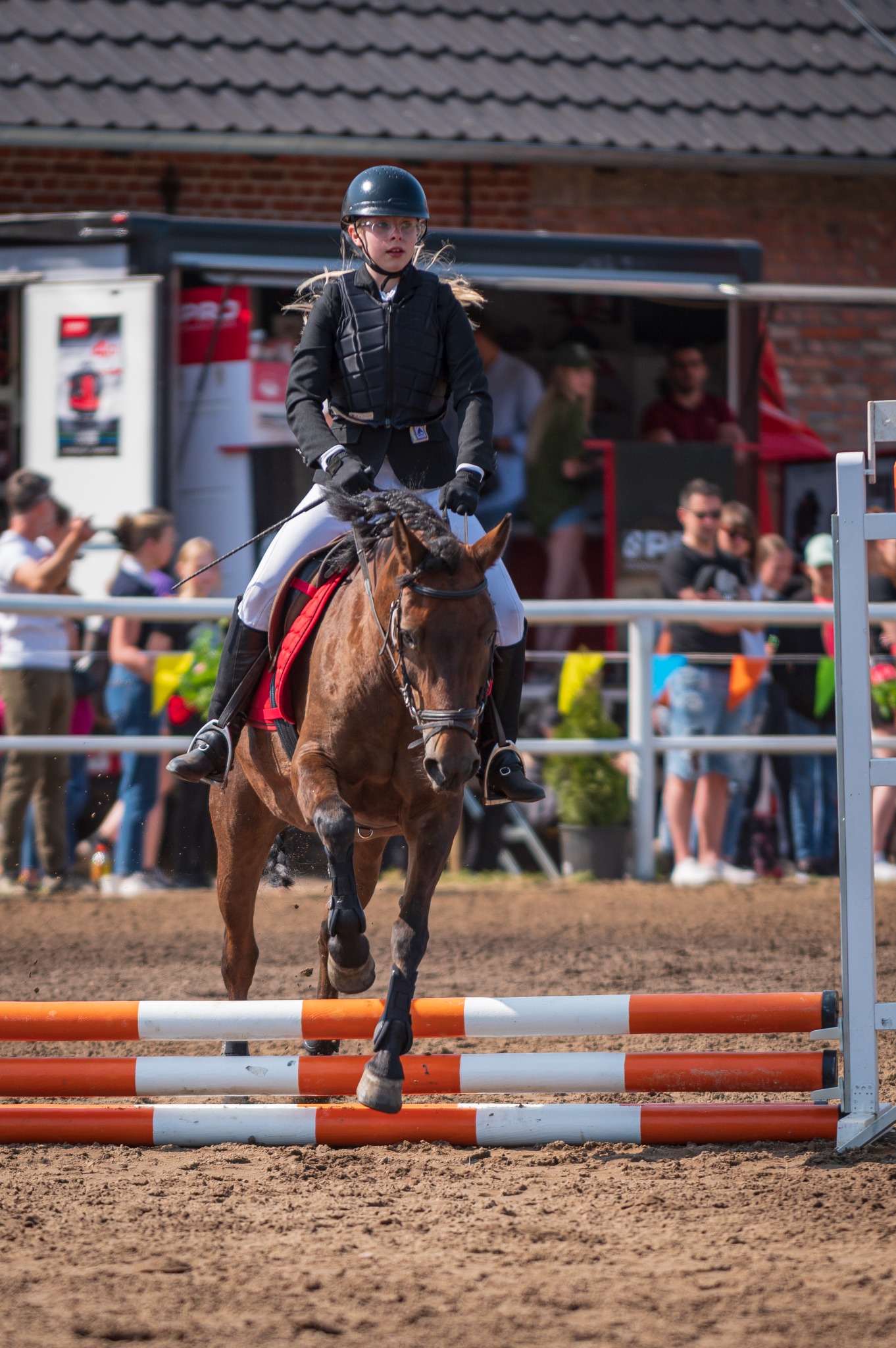 Młoda amazonka w czarnym kasku i kamizelce, na brązowym koniu skacze przez przeszkodę z pomarańczowo-białych drągów na zawodach jeździeckich, w tle widoczni kibice i budynki.