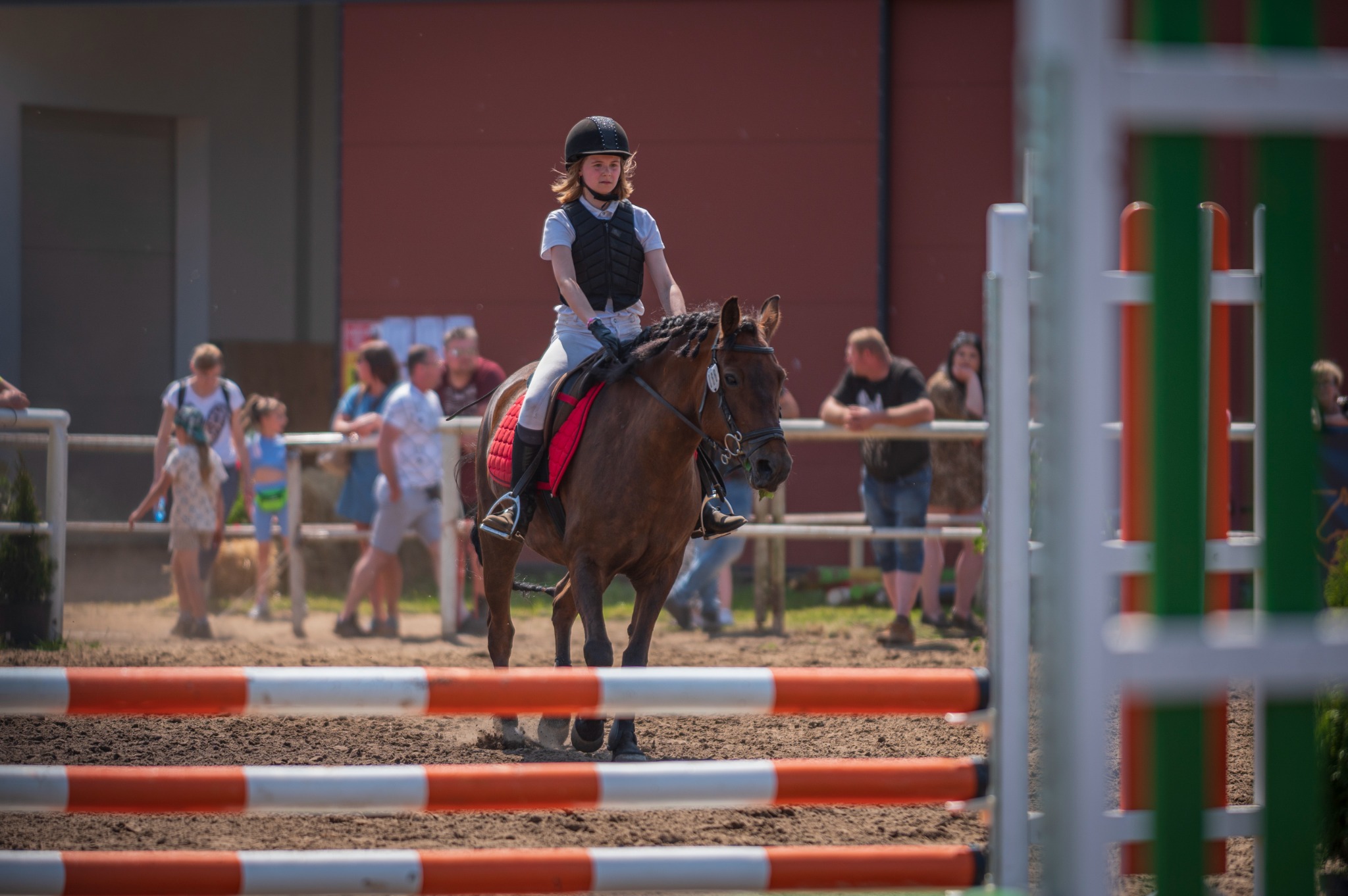 Młoda dziewczyna w czarnym kasku i kamizelce jeździeckiej na brązowym koniu pokonuje przeszkodę na torze, widzowie w tle.