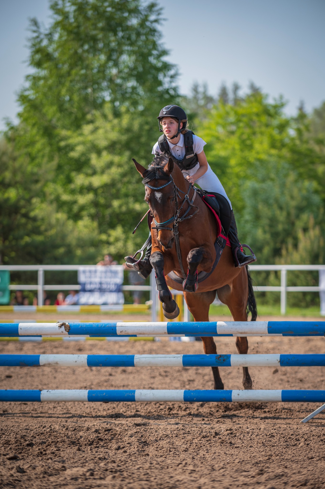 Młoda amazonka w kasku i kamizelce ochronnej skacze na brązowym koniu przez przeszkodę z niebiesko-białych drągów na piaszczystym terenie.