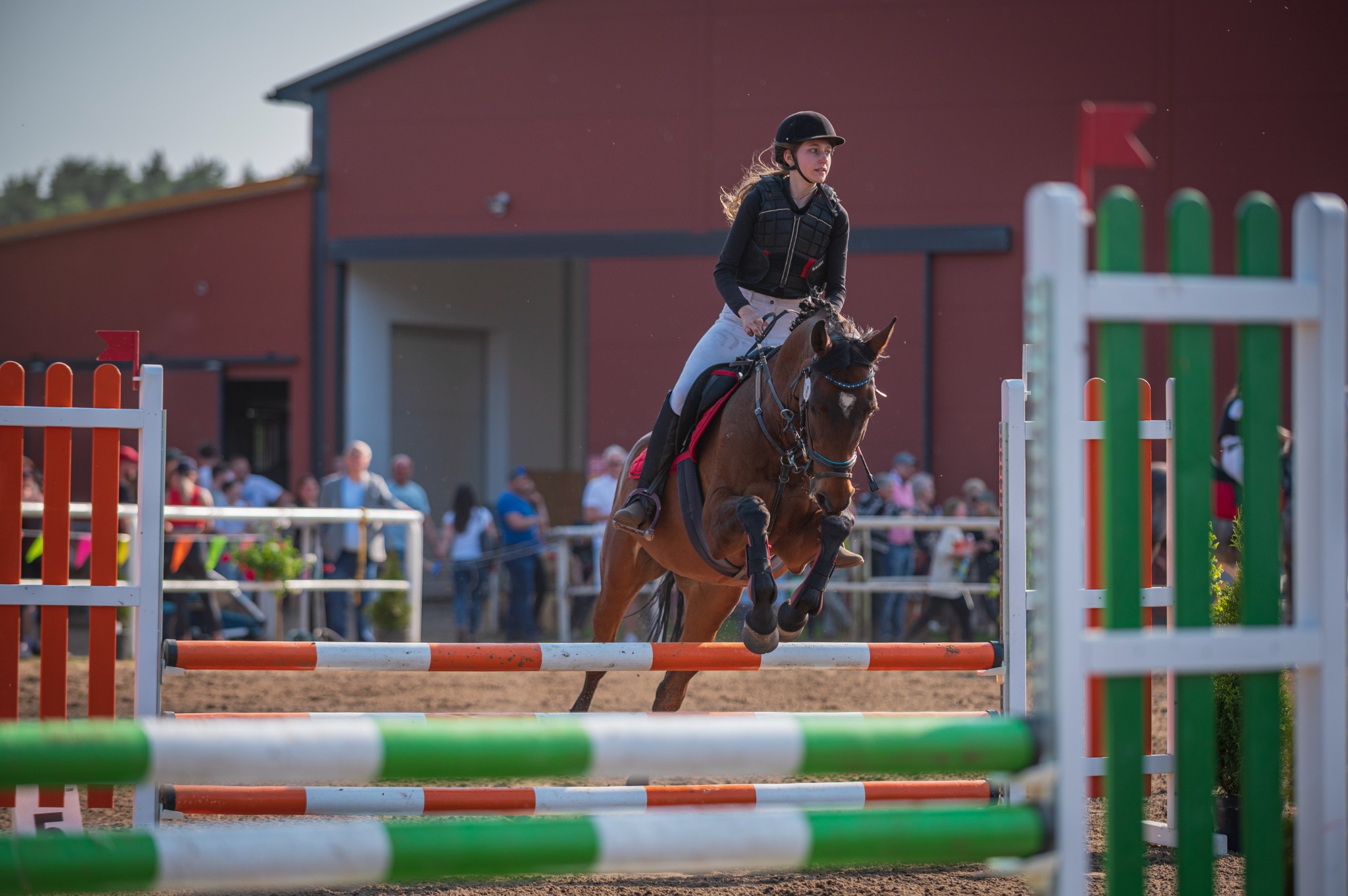 Młoda kobieta w kamizelce ochronnej i kasku skacze na brązowym koniu przez przeszkodę z pomalowanymi na pomarańczowo-biało i zielono-biało poprzeczkami, w tle widoczny budynek i publiczność.