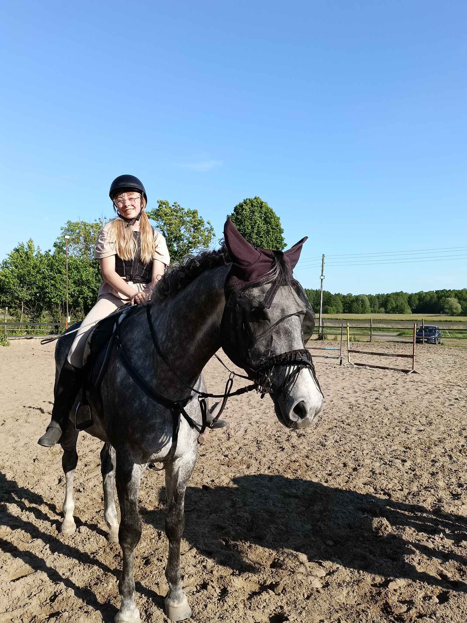 Uśmiechnięta dziewczyna w czarnym kasku i kamizelce ochronnej siedzi na siwym koniu z maską na pysku na piaszczystym terenie, w tle widoczne drzewa i błękitne niebo.