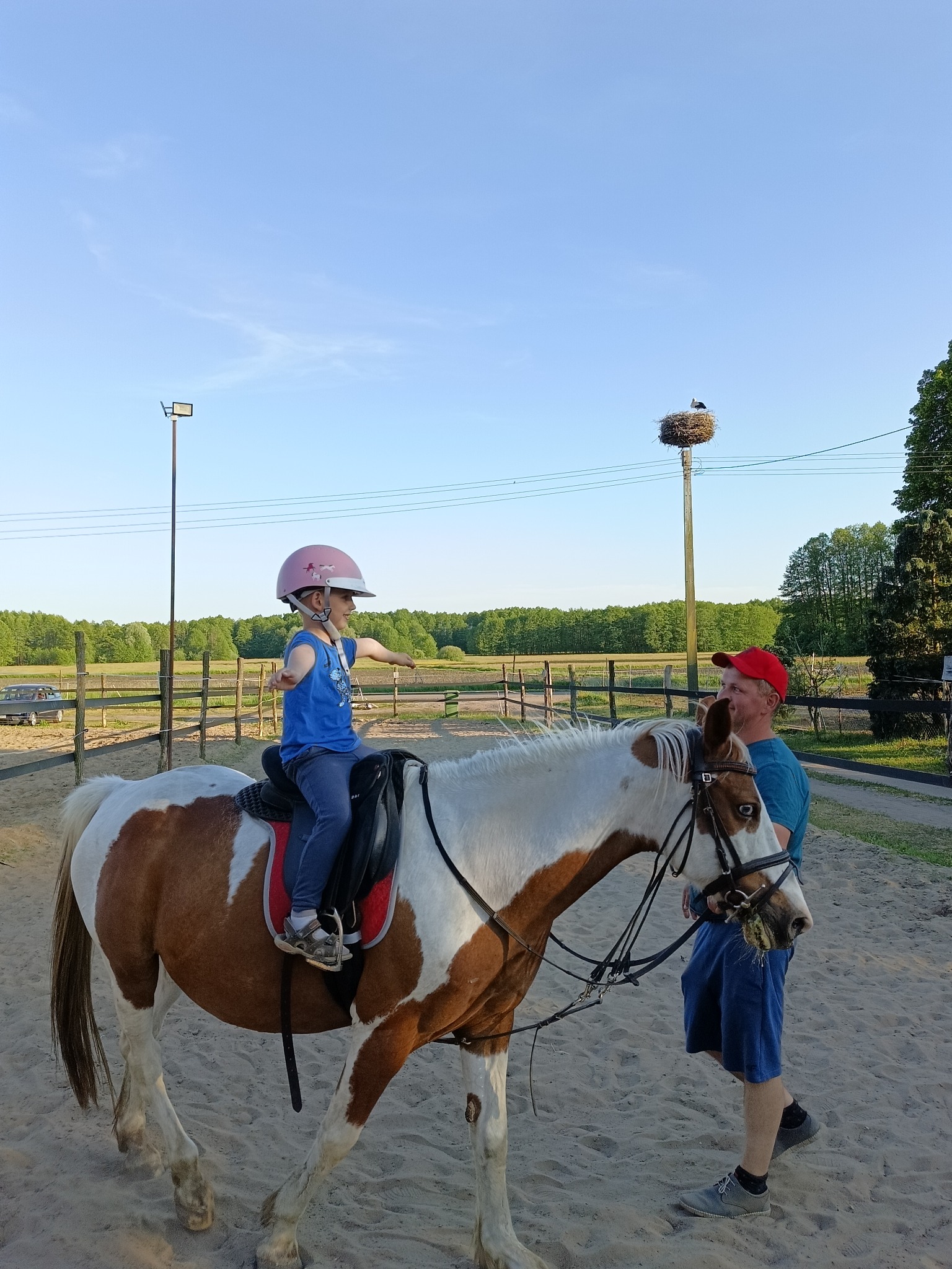 Dziewczynka w różowym kasku uczy się jeździć na brązowo-białym koniu pod okiem instruktora, w tle widoczny słup z gniazdem bocianim.