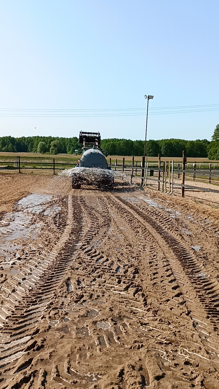 Ciężki traktor z cysterną rozpyla wodę na błotnistej nawierzchni placu treningowego, tworząc głębokie ślady opon, z widocznym lasem i lampą oświetleniową w tle.