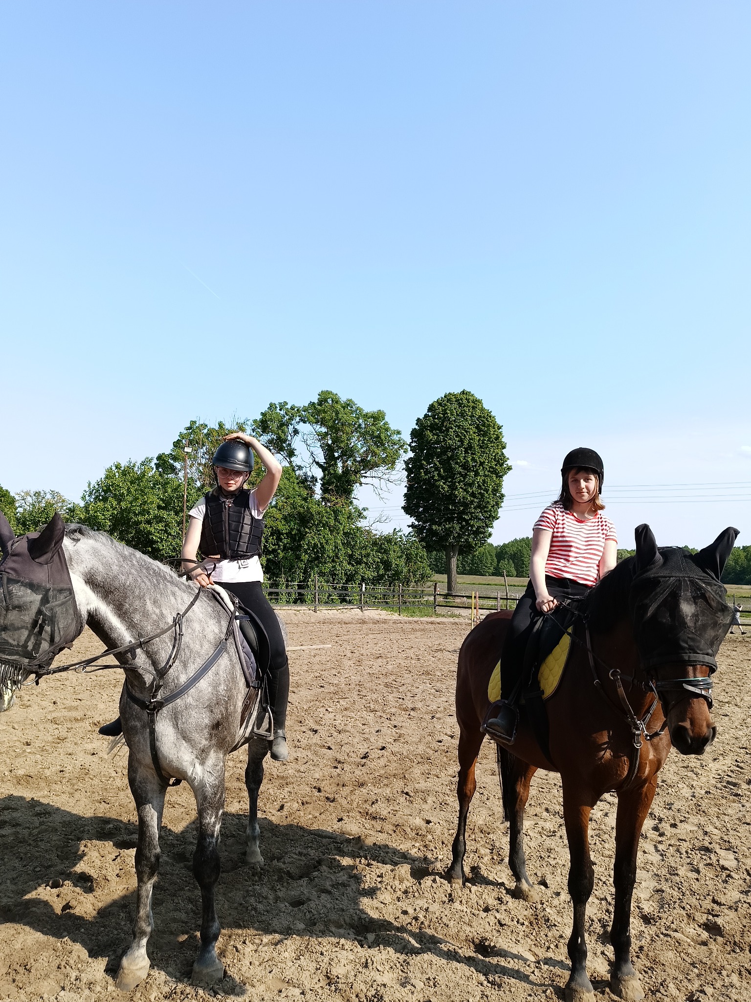 Dwie osoby na koniach na piaszczystym terenie, jedna z nich poprawia kask, konie mają nałożone siatki na głowy.