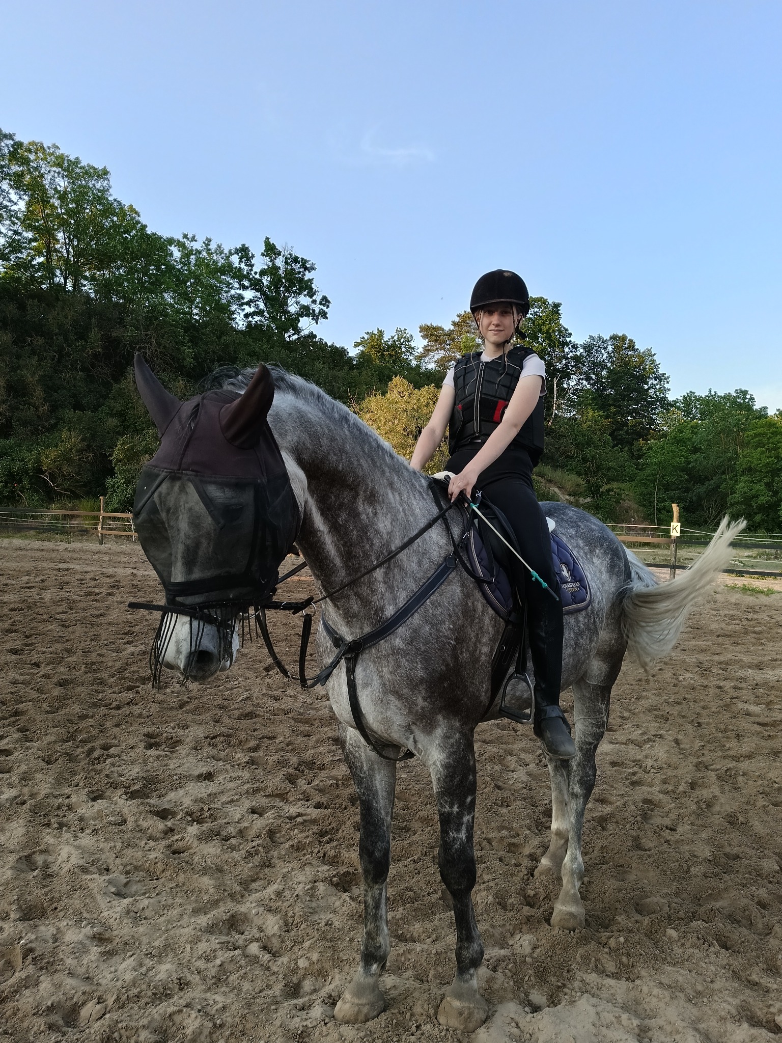 Dziewczyna w kamizelce ochronnej i kasku siedzi na siwym koniu z maską na głowie na piaszczystym terenie.