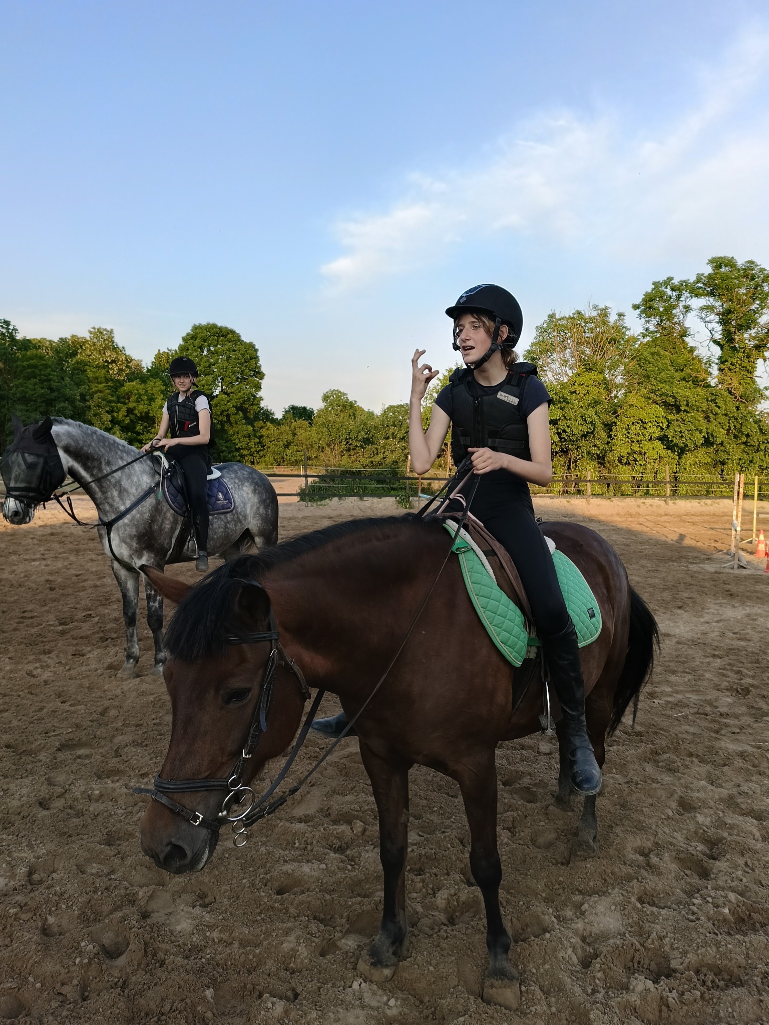 Dwie osoby na koniach na piaszczystym terenie, jedna z nich gestykuluje dłonią, obie w kaskach i kamizelkach ochronnych.