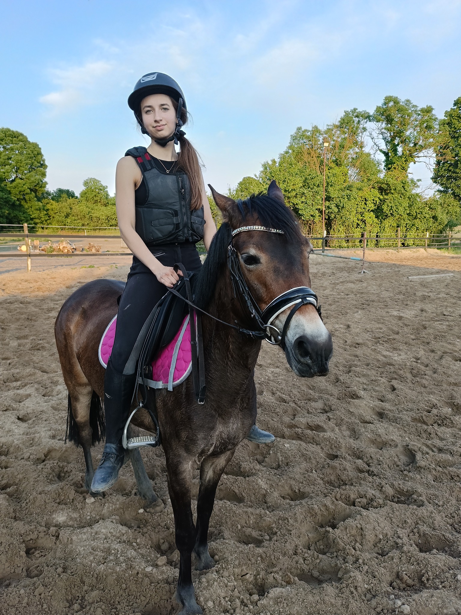 Młoda kobieta w kasku i kamizelce ochronnej siedzi na brązowym koniu z ozdobnym ogłowiem na piaszczystym padoku.