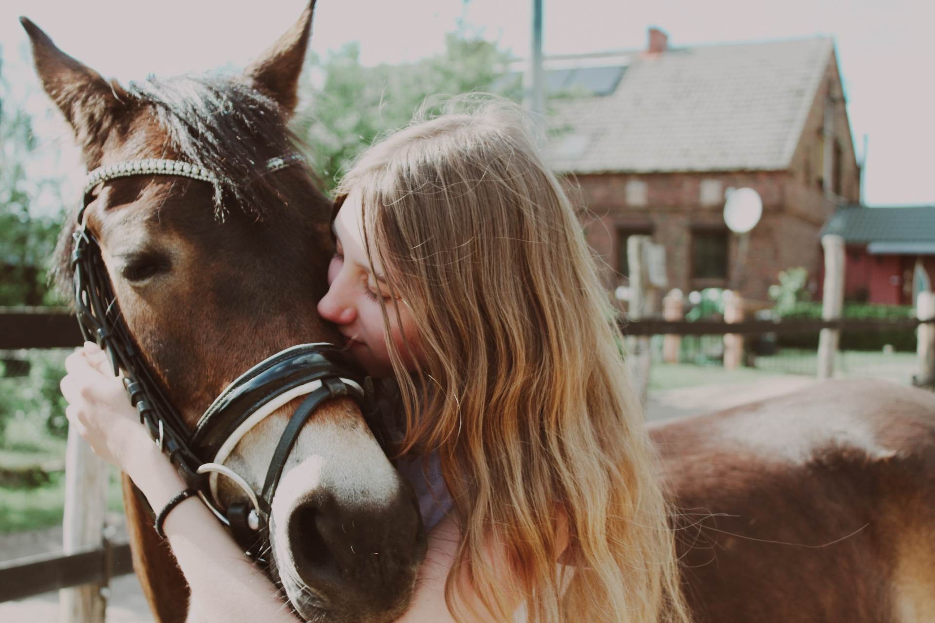 Młoda kobieta obejmuje brązowego konia z uprzężą ozdobioną kryształkami, na tle zabudowań gospodarskich