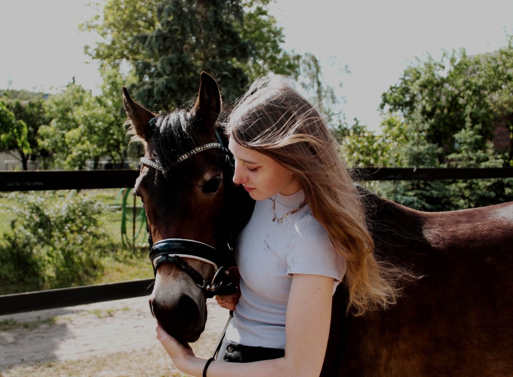 Młoda kobieta z długimi włosami obejmuje brązowego konia z ozdobnym ogłowiem, na tle zieleni i ogrodzenia.