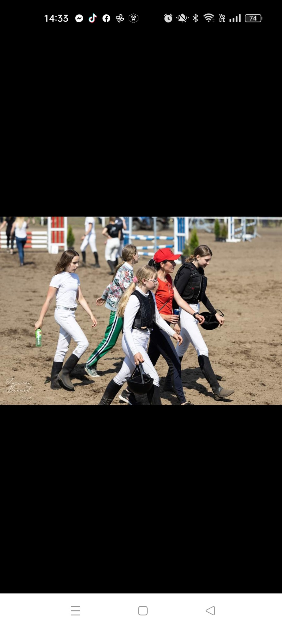 Grupa młodych osób w strojach jeździeckich, niektóre z kaskami, przechodzi przez piaszczysty teren w pobliżu przeszkód do skoków.