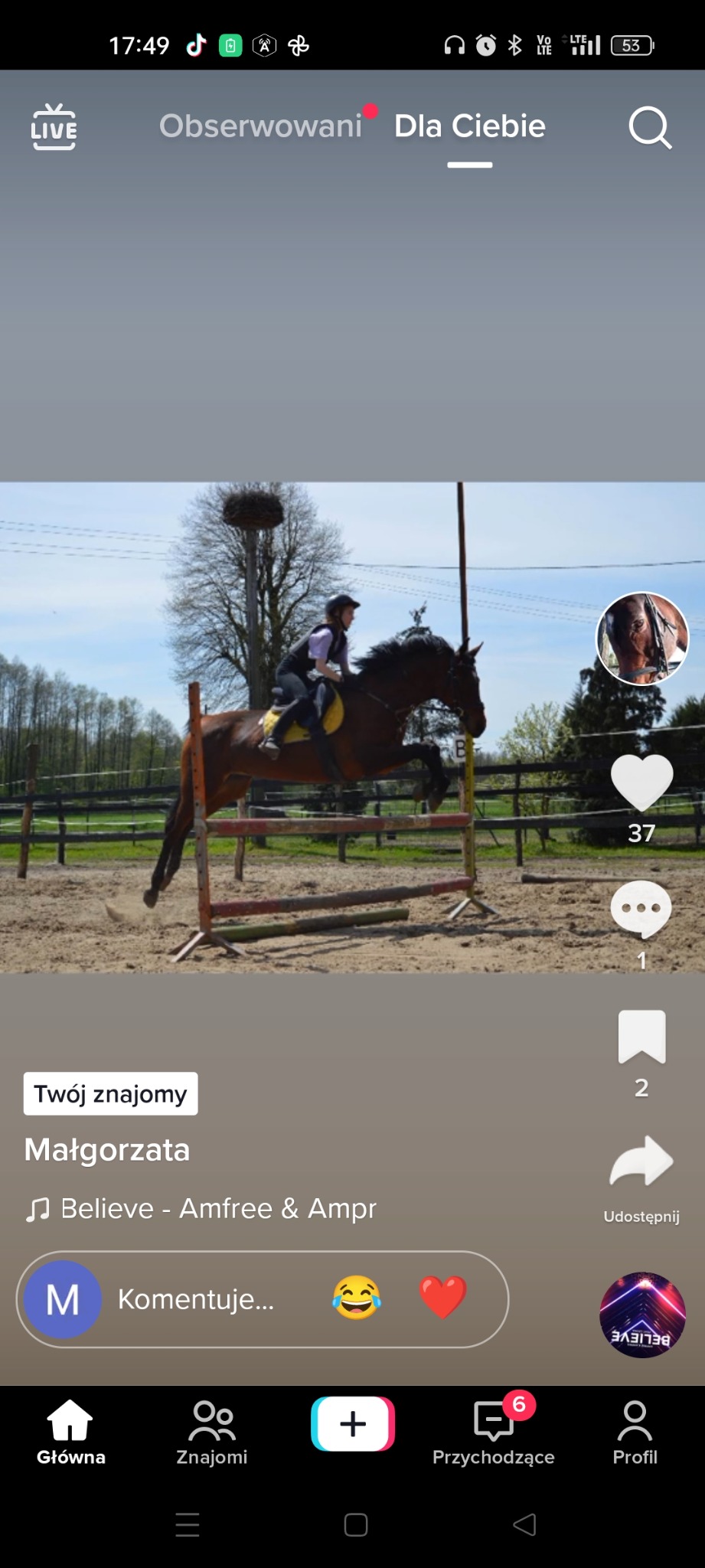 Jeździec na brązowym koniu skacze przez przeszkodę na piaszczystym terenie, widoczne ogrodzenie i drzewa w tle, zdjęcie zrobione z perspektywy ekranu telefonu.