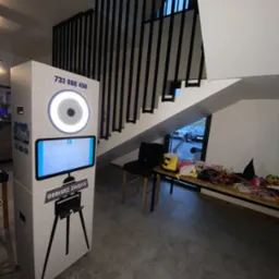 Fotobudka z oświetleniem pierścieniowym i ekranem dotykowym, obok stolik z rekwizytami do zdjęć i schody z czarną balustradą.