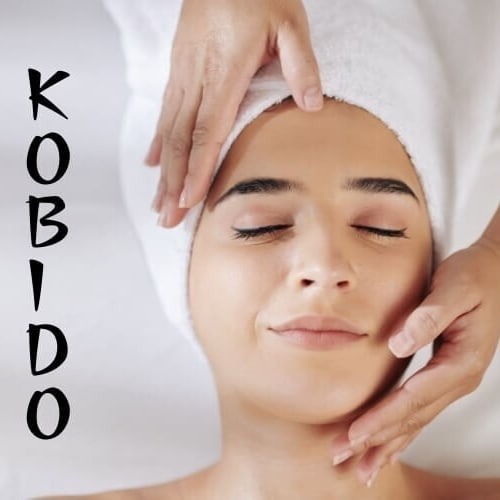 Kobieta z zamkniętymi oczami, owinięta białym ręcznikiem na głowie, podczas masażu Kobido w Lublinie. Dłonie masażysty delikatnie dotykają jej twarzy.