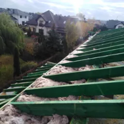 Zielona drewniana konstrukcja dachu z izolacją z wełny mineralnej, widok na domy jednorodzinne w tle.