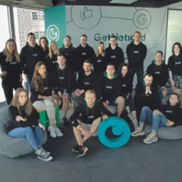 Zespół firmy 'Get Noticed' w czarnych bluzach z zielonymi akcentami, pozujący na tle biura z logo i widokiem na miasto, z dużym, turkusowym logo firmy na pierwszym planie.