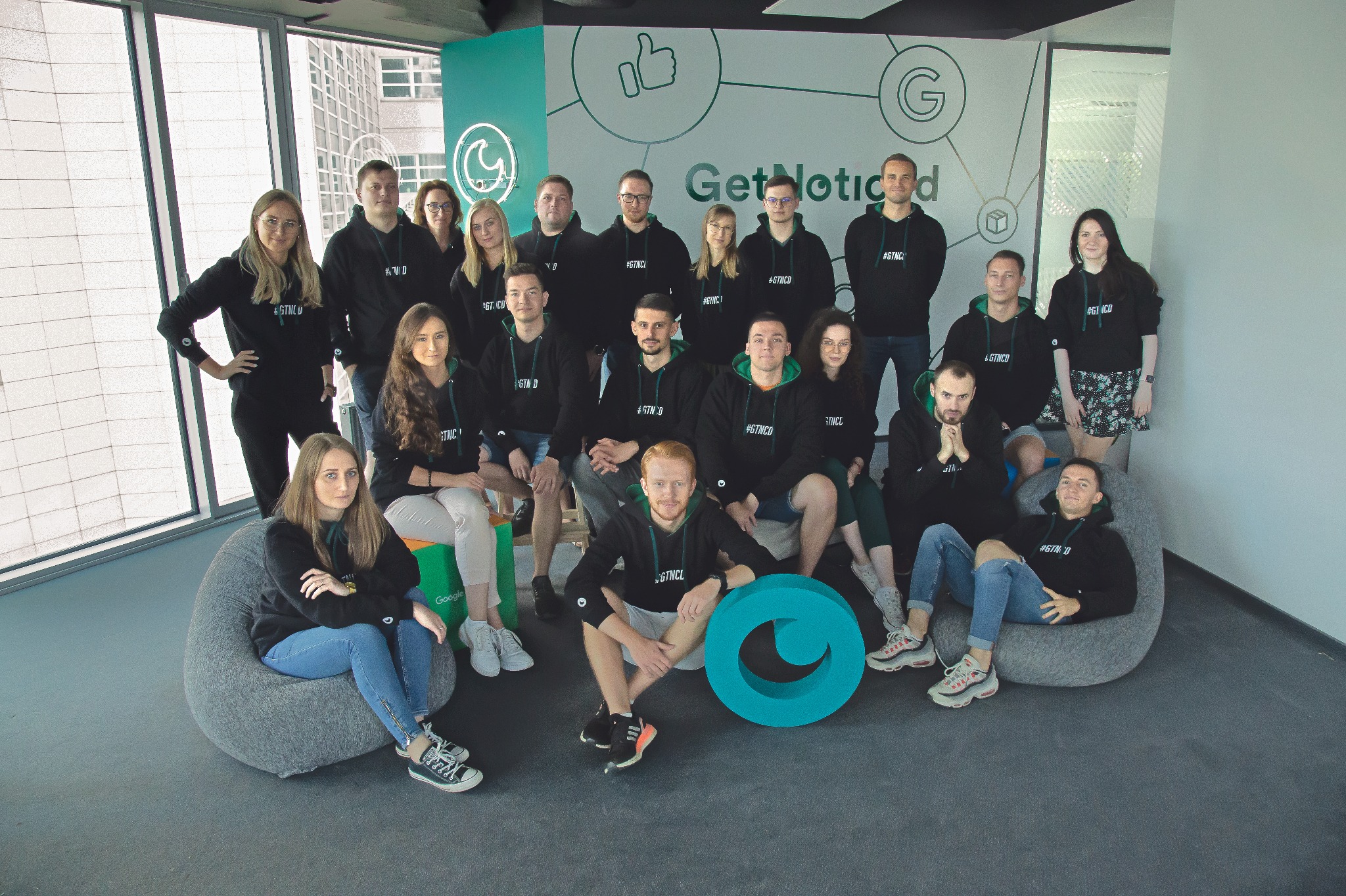 Zespół firmy 'Get Noticed' w czarnych bluzach z zielonymi akcentami, pozujący na tle biura z logo i widokiem na miasto, z dużym, turkusowym logo firmy na pierwszym planie.