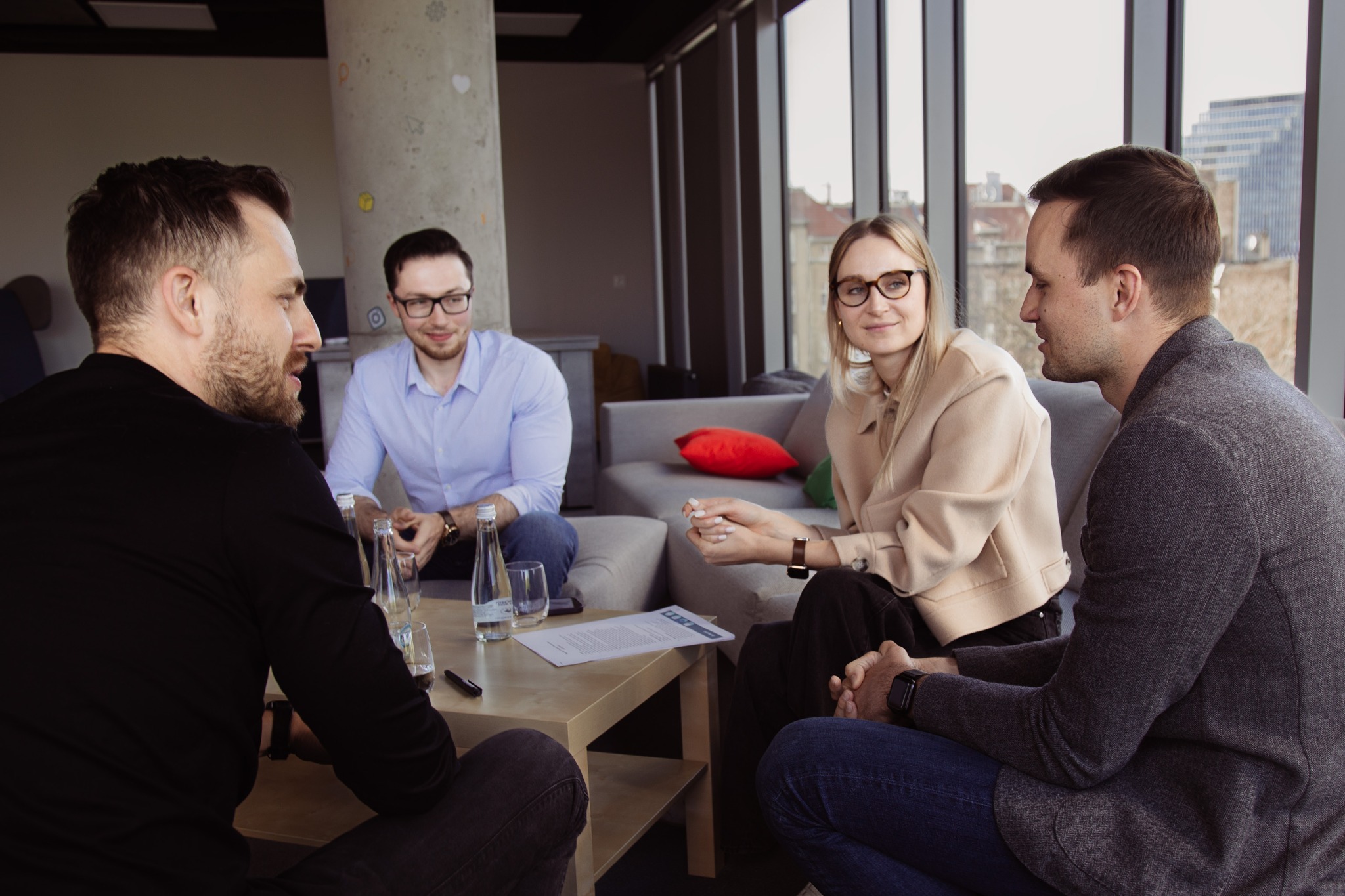 Cztery osoby siedzą przy stoliku podczas spotkania w nowoczesnym biurze z betonowym filarem i widokiem na miasto za oknem.