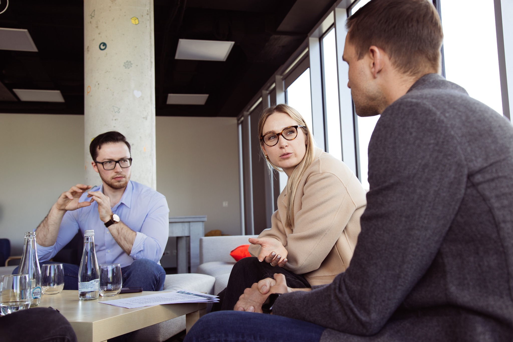 Trzech specjalistów w trakcie spotkania w nowoczesnym biurze z ozdobionym filarem, omawiających dokumenty leżące na stole.