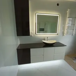 Nowoczesna łazienka z podświetlanym lustrem, umywalką nablatową w kształcie łódki i szafką z czarno-białymi frontami oraz wysoką, wiszącą szafką w kolorze czarnym.