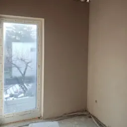 Świeżo nałożony tynk maszynowy na ścianie wewnątrz budynku, widoczne przewody instalacyjne przy podłodze i okno PCV z widokiem na zewnątrz.