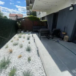 Nowoczesny balkon z szarymi płytkami i białym żwirem, roślinami, oświetleniem girlandą, meblami ogrodowymi i zielonym żywopłotem w tle.