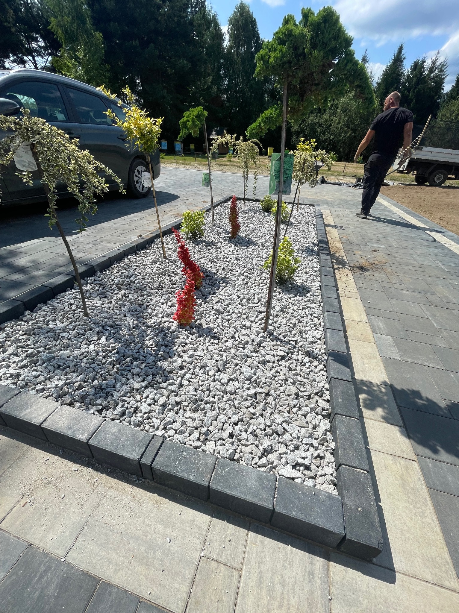 Obrzeża z kostki brukowej okalają rabatę z kamykami i roślinami. Ułożona nawierzchnia z kostki w odcieniach szarości. Prace wykończeniowe w ogrodzie, widoczny pracownik.