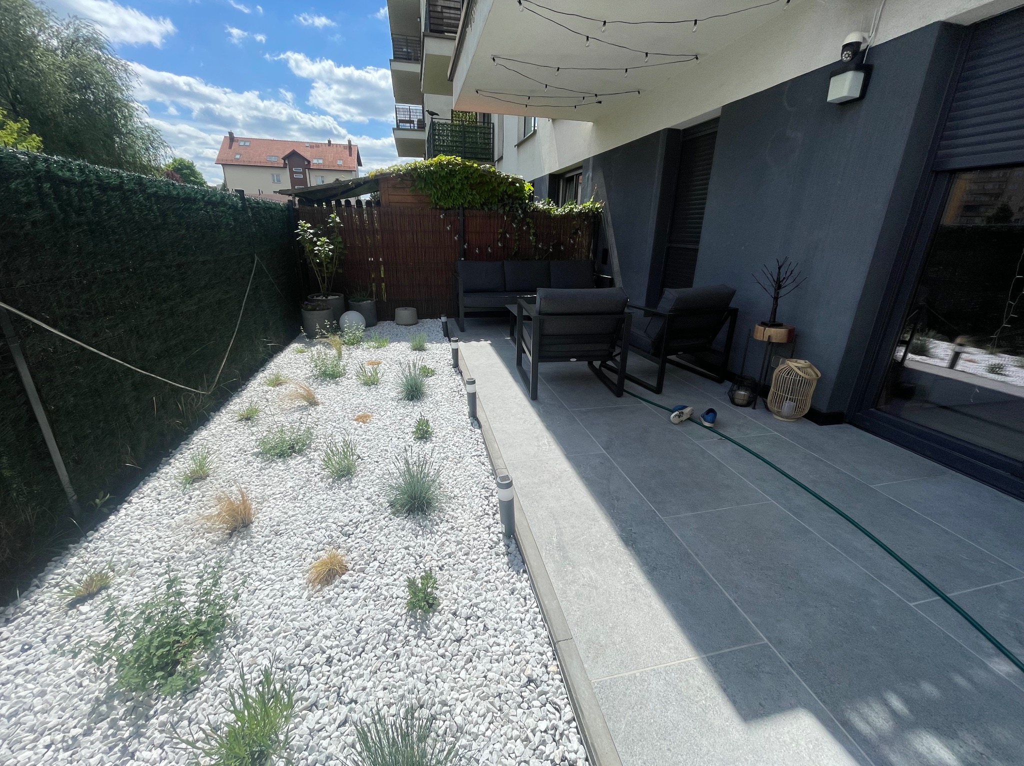 Nowoczesny balkon z szarymi płytkami i białym żwirem, roślinami, oświetleniem girlandą, meblami ogrodowymi i zielonym żywopłotem w tle.