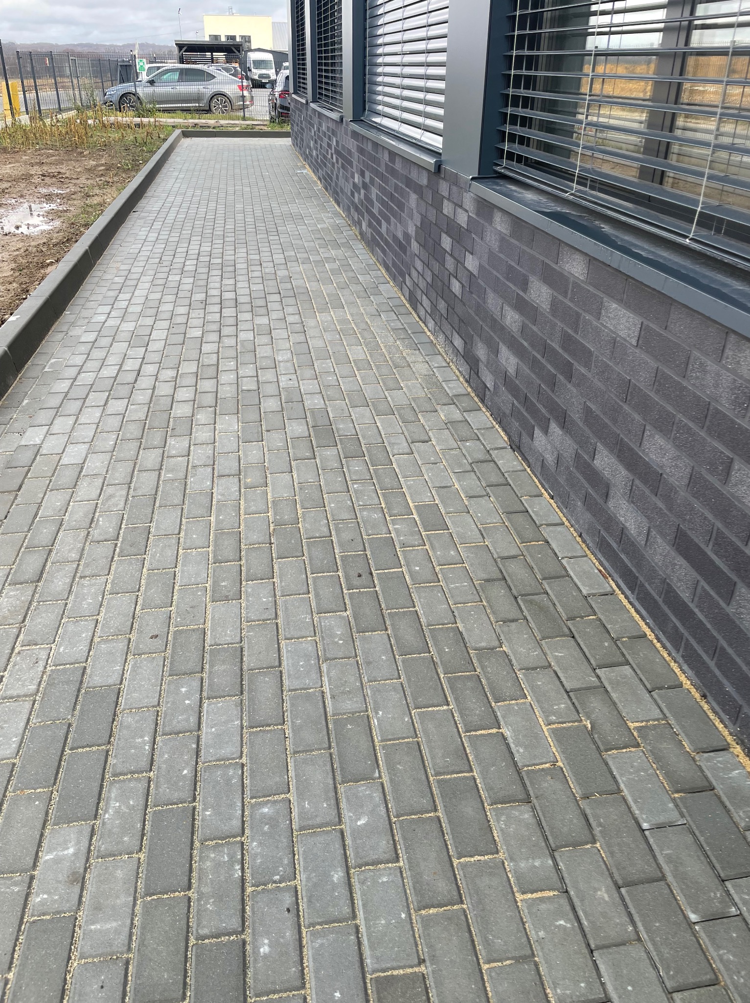 Szara kostka brukowa ułożona wzdłuż budynku z ciemnej cegły. Widoczne obrzeża i samochód w tle. Nowoczesna nawierzchnia.
