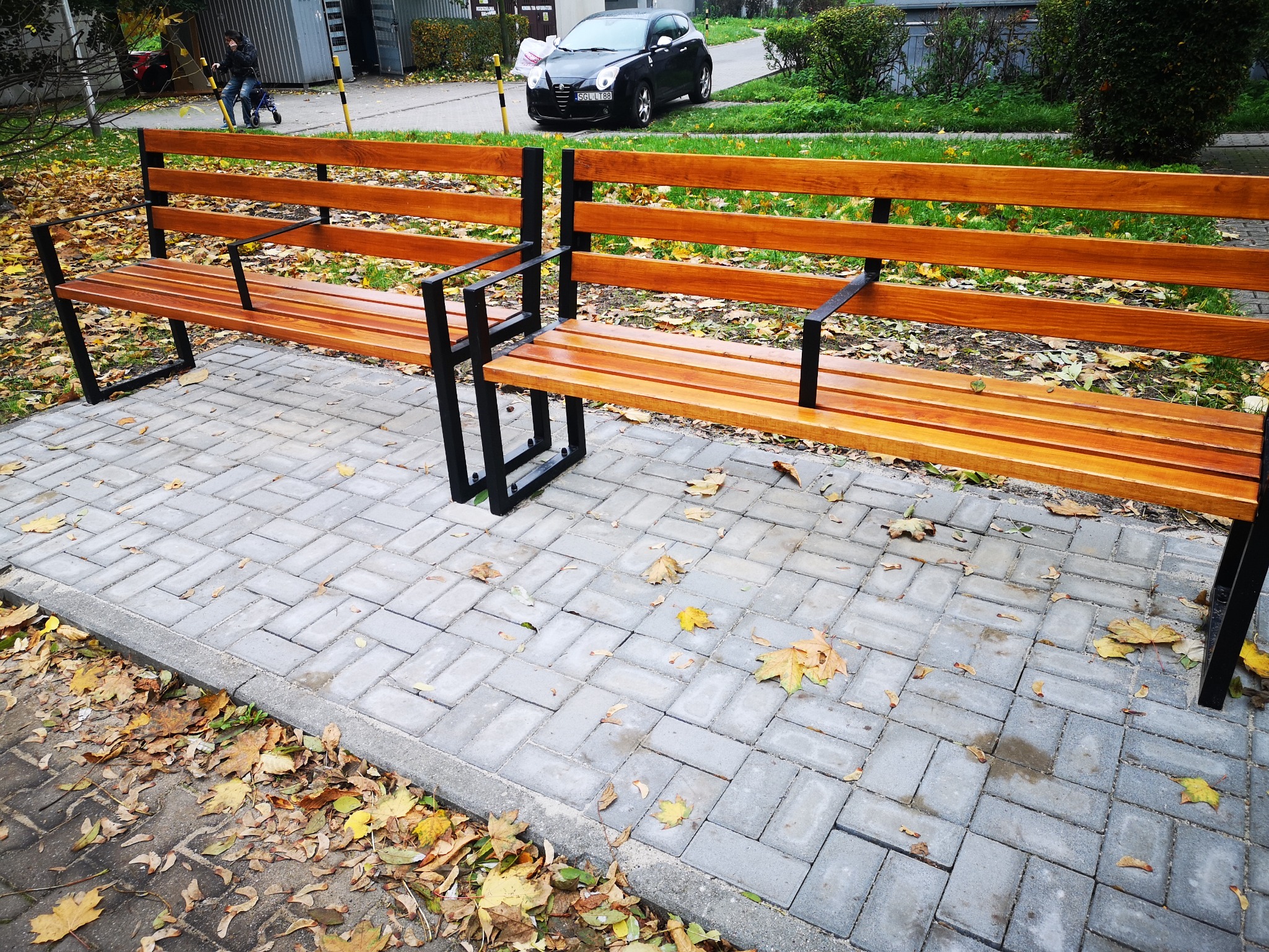 Dwie drewniane ławki z czarnymi metalowymi nogami stojące na szarej kostce brukowej, otoczone opadłymi liśćmi. W tle widoczny fragment ulicy z zaparkowanym czarnym samochodem oraz przechodzący...