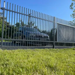 Szara, metalowa brama wjazdowa z pionowymi szczebelkami, za którą widać samochód osobowy, zieleń drzew i błękitne niebo.