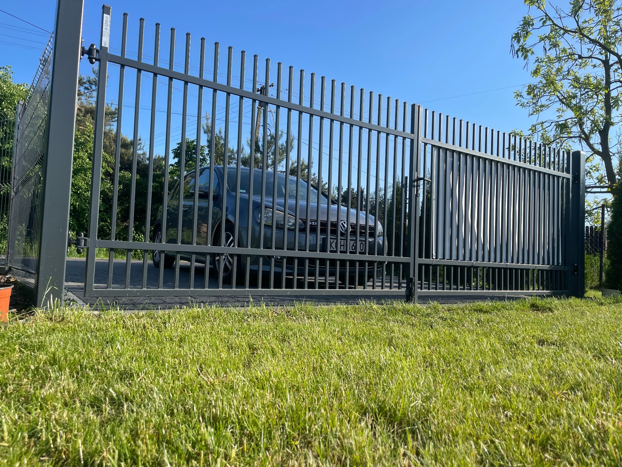 Szara, metalowa brama wjazdowa z pionowymi szczebelkami, za którą widać samochód osobowy, zieleń drzew i błękitne niebo.