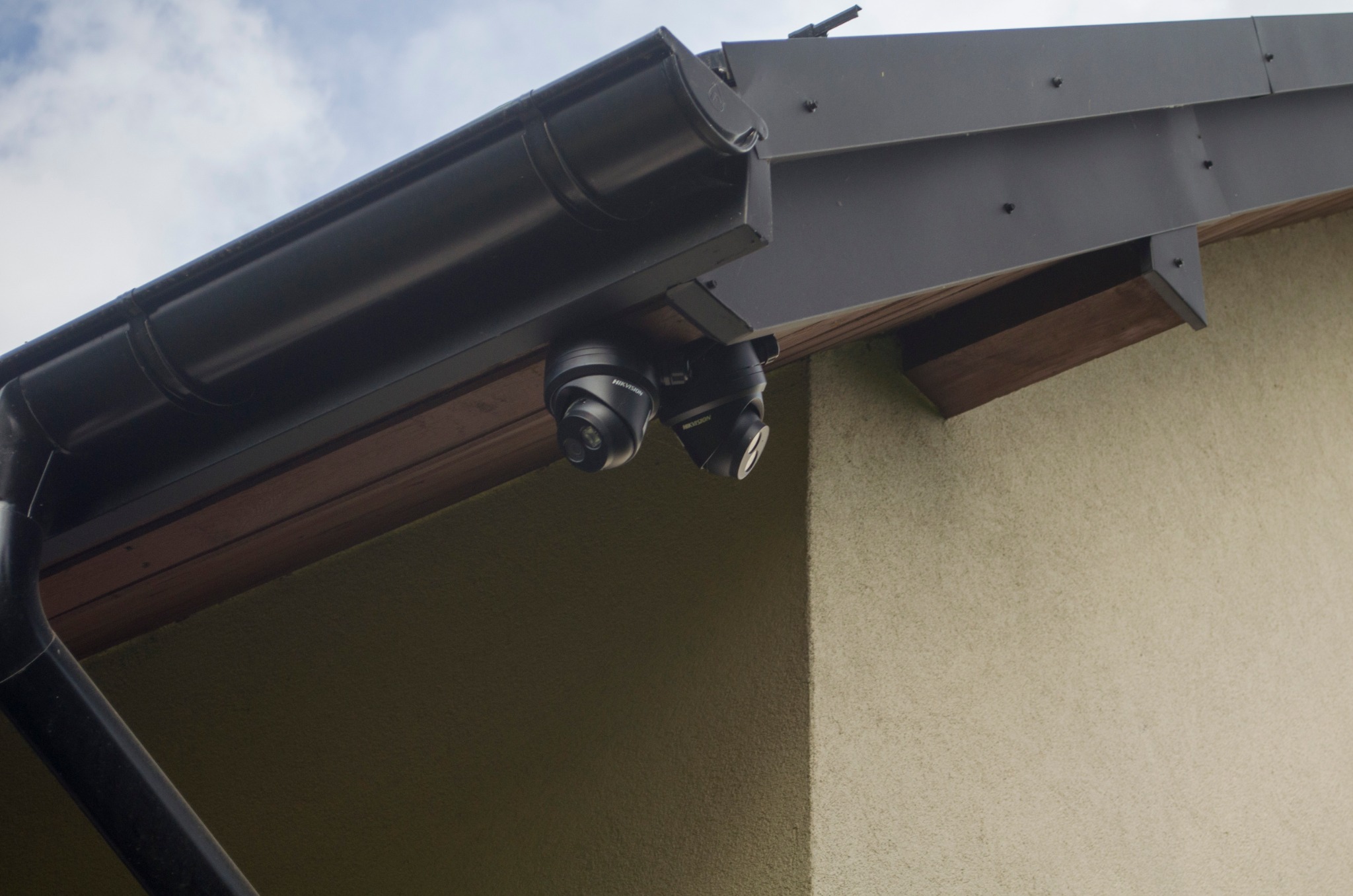 Dwie czarne kamery kopułkowe marki Hikvision zamontowane pod okapem dachu, widoczne rynna i fragment elewacji budynku.