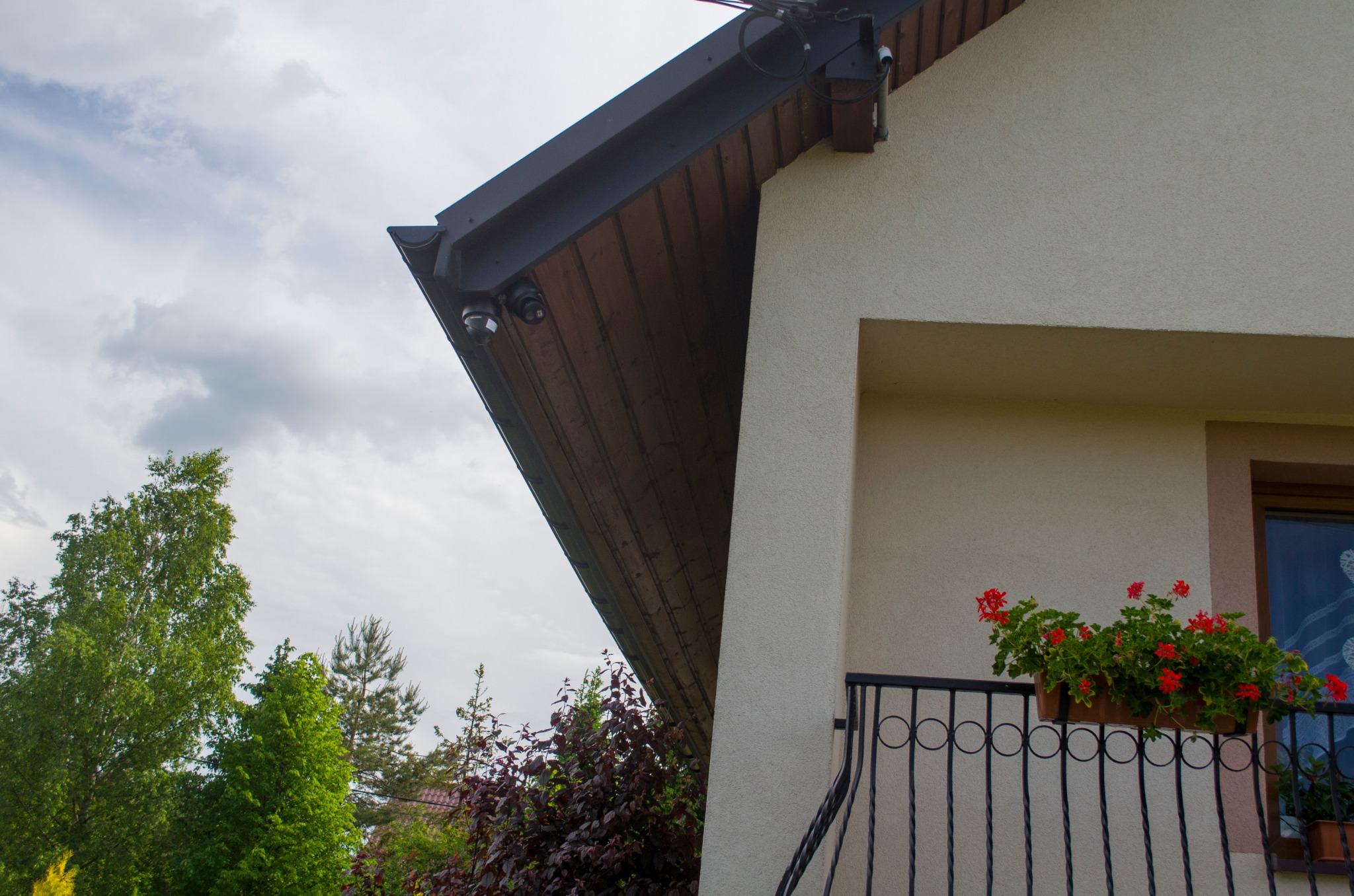 Dwie kamery monitoringu zamontowane pod okapem domu z balkonem i kwiatami w doniczce, widok z dołu na tle zieleni i pochmurnego nieba.