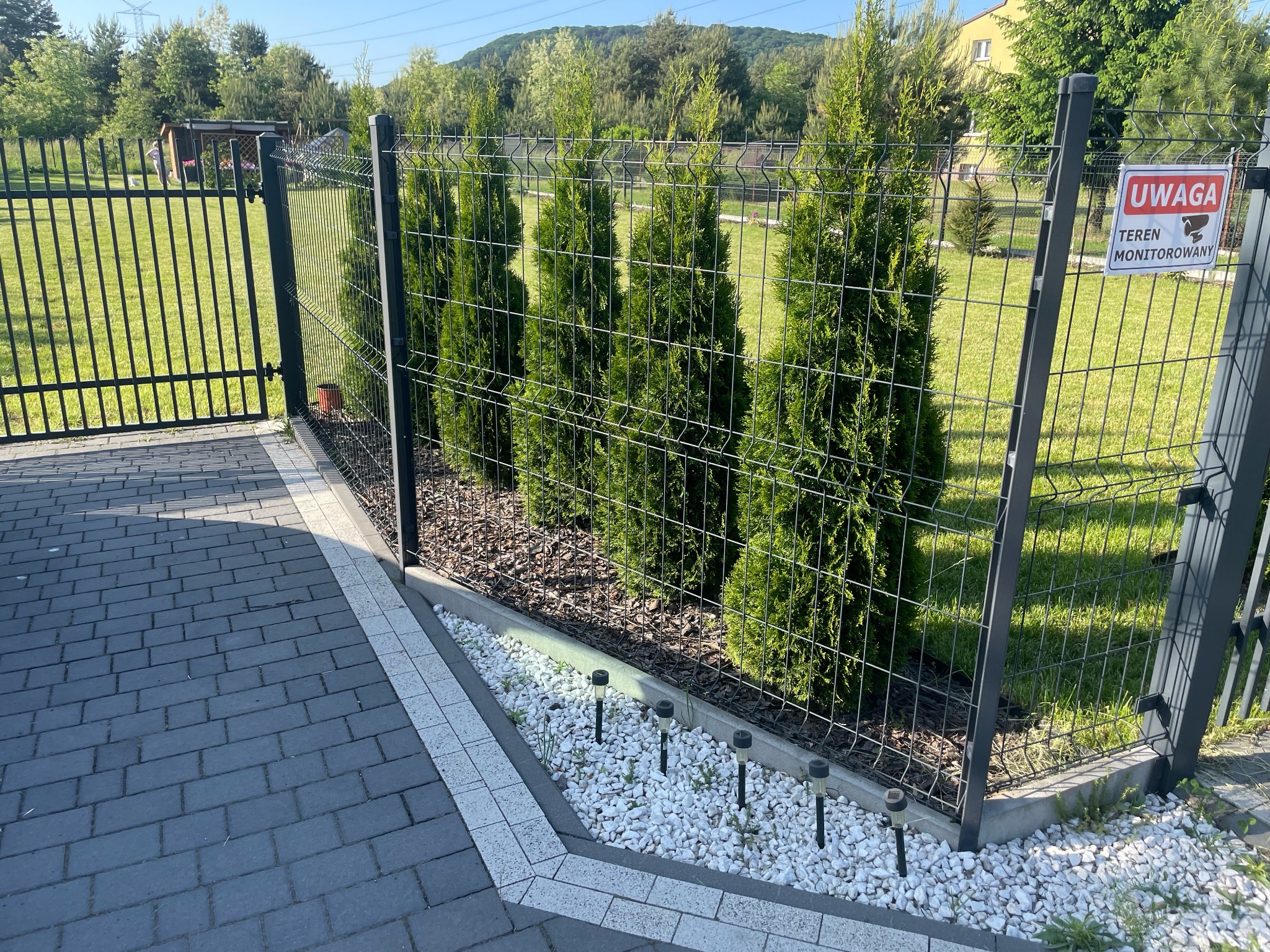 Nowoczesne ogrodzenie panelowe w Libiążu z nasadzeniami tuj i oświetleniem punktowym, widoczny znak ostrzegawczy o monitoringu.