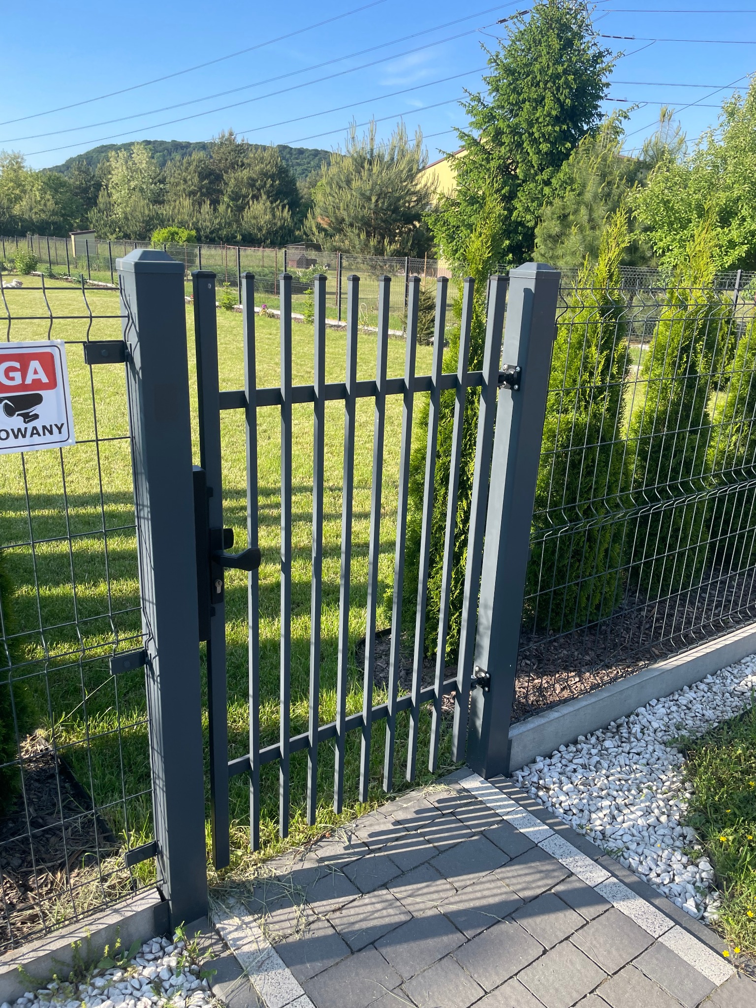 Grafitowa furtka z pionowymi szczebelkami, część ogrodzenia panelowego z siatki, widoczny znak ostrzegawczy z kamerą, kostka brukowa i białe kamienie ozdobne, w tle zieleń ogrodu i drzewa.