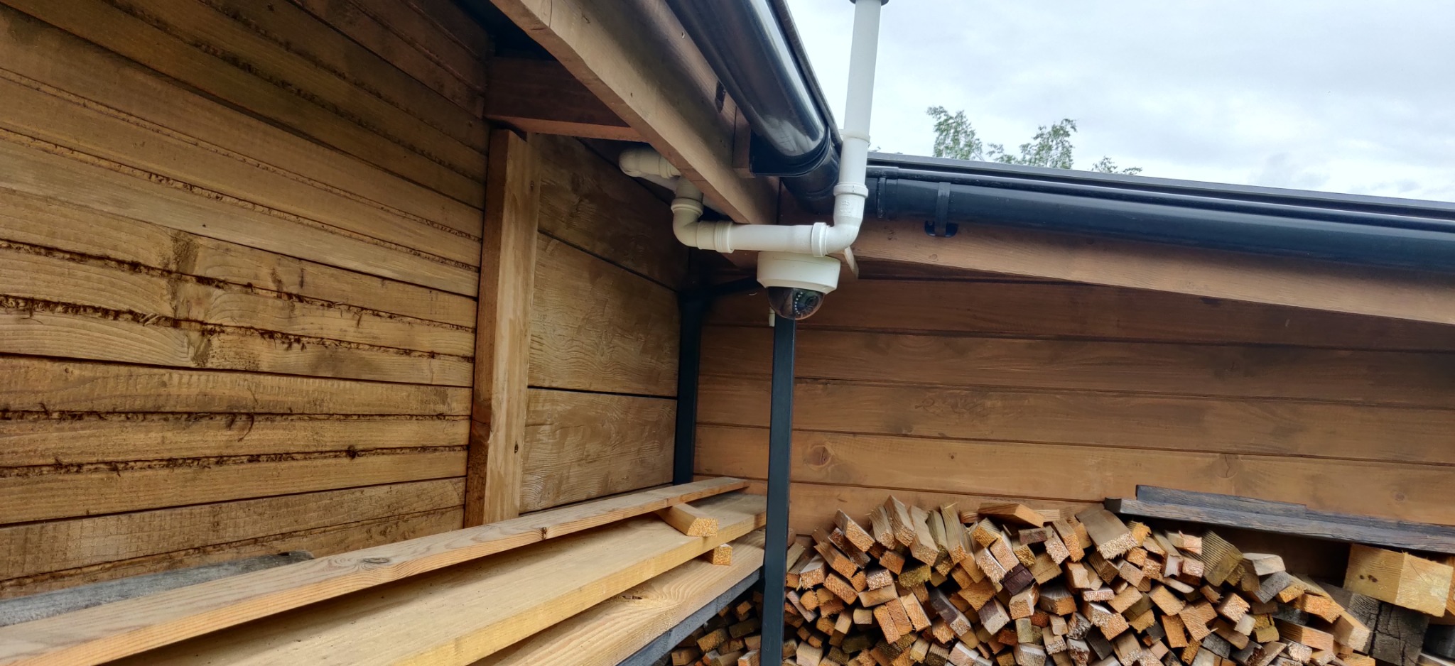 Kamera monitoringu kopułkowa zamontowana pod okapem drewnianej konstrukcji, widoczne rury instalacyjne i stos drewna.