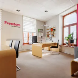 Jasne biuro agencji nieruchomości z nowoczesnymi meblami, r&oacute;żową ścianą akcentującą i logo 'Freedom nieruchomości' nad oknem.